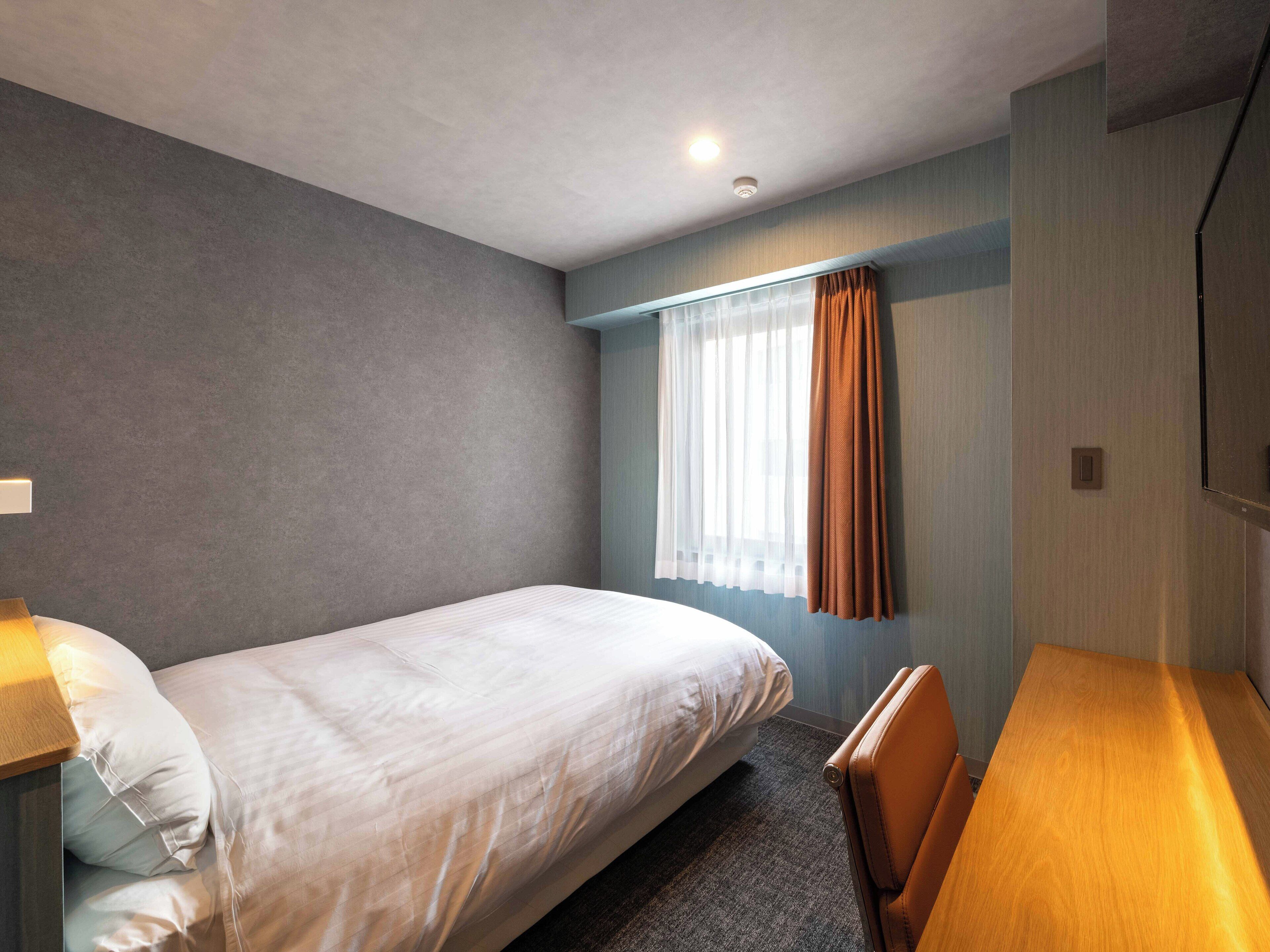 A single bed with a headboard, a wooden desk, a chair, a window with sheer curtains, and a wall-mounted light fixture.