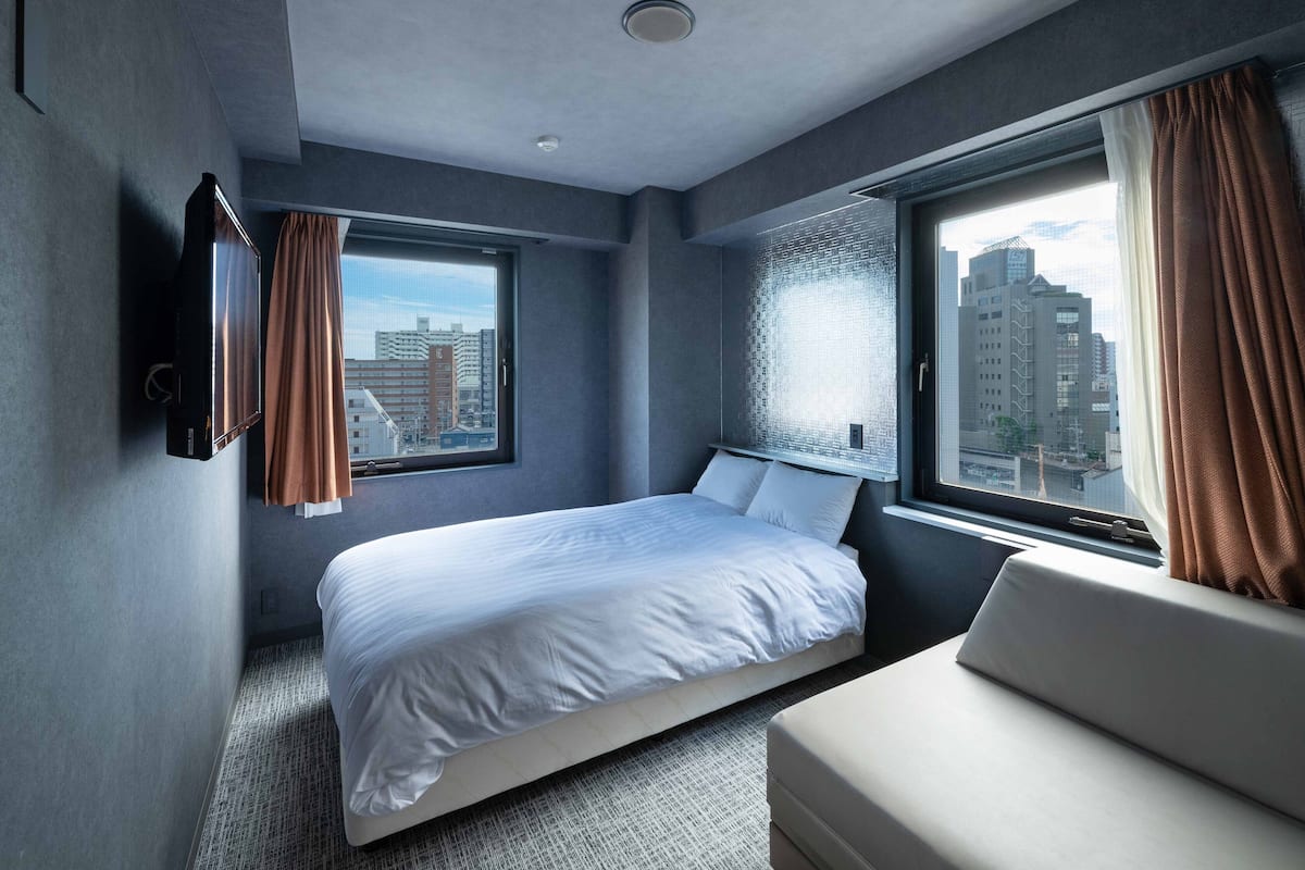 A modern hotel room with a large bed, a sofa, and a city view through the windows.