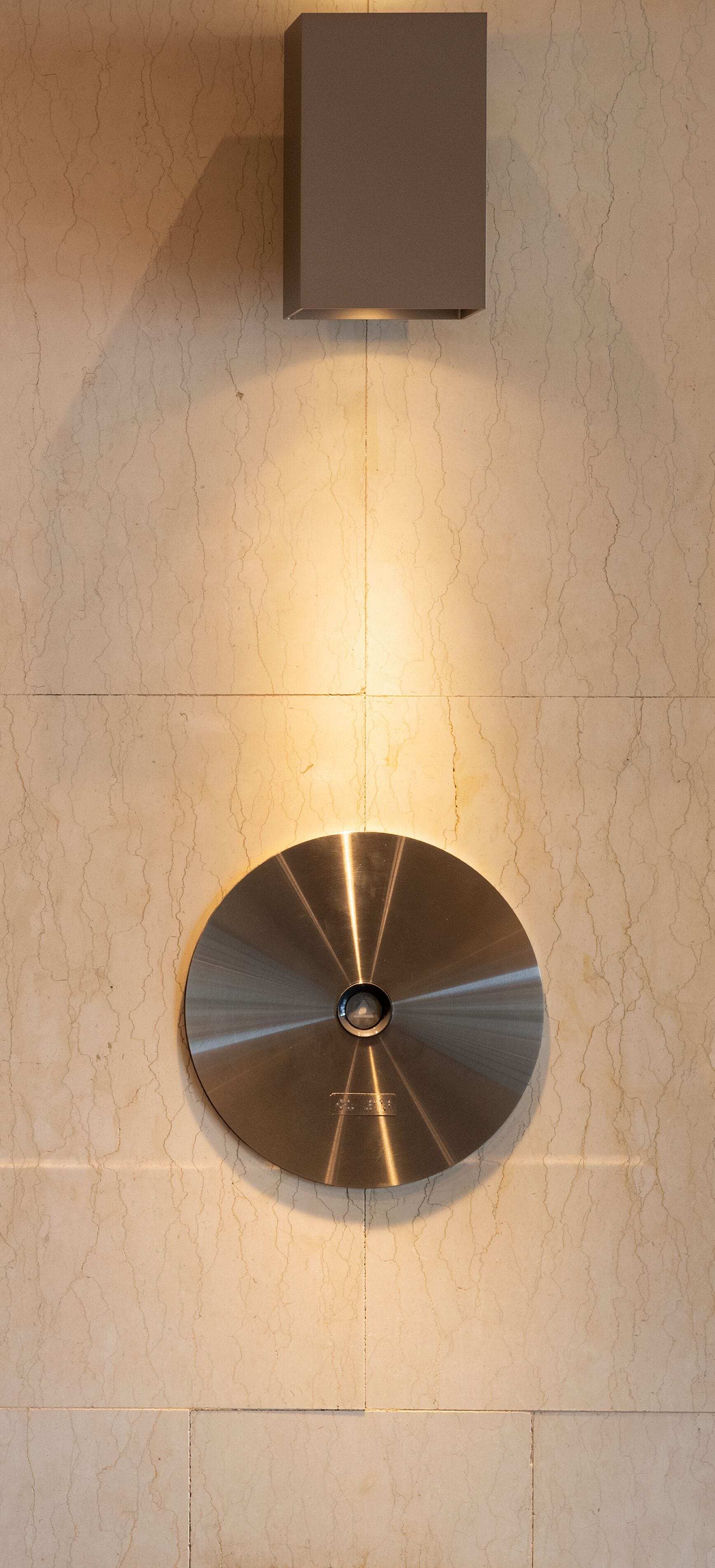 A wall-mounted, circular metallic object with a central hole, likely a decorative or functional piece, against a textured wall with a square object above it.