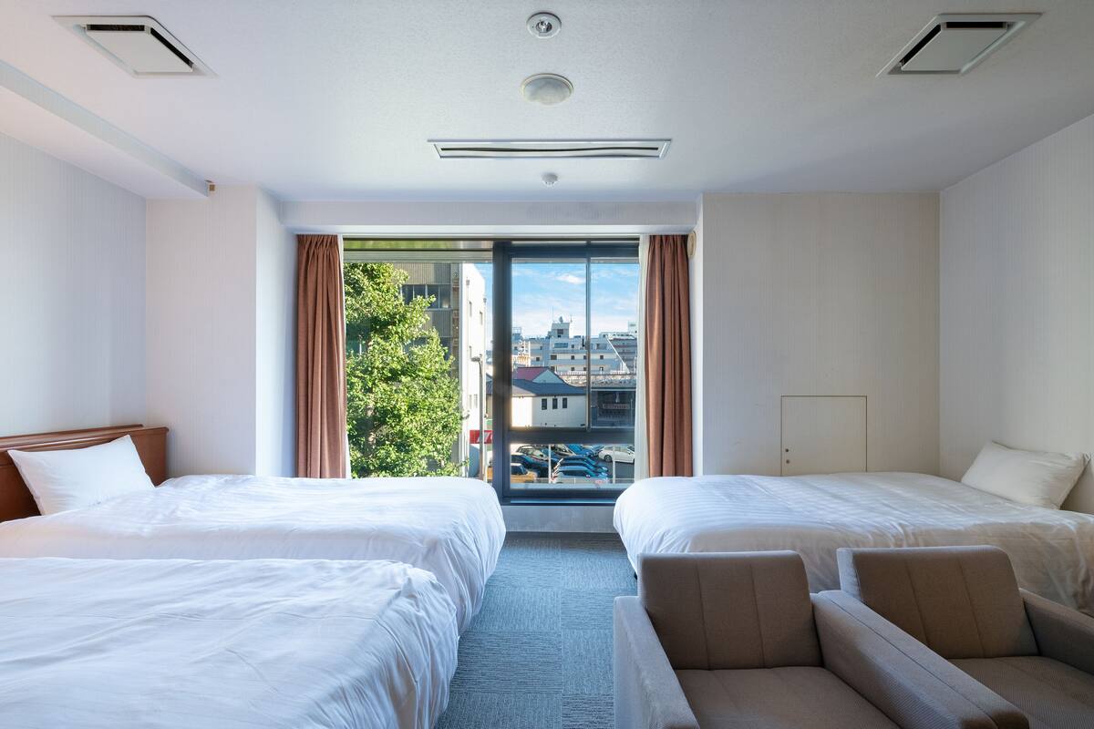 A hotel room with two beds, a sofa, a large window with curtains, and a view of buildings and trees outside.