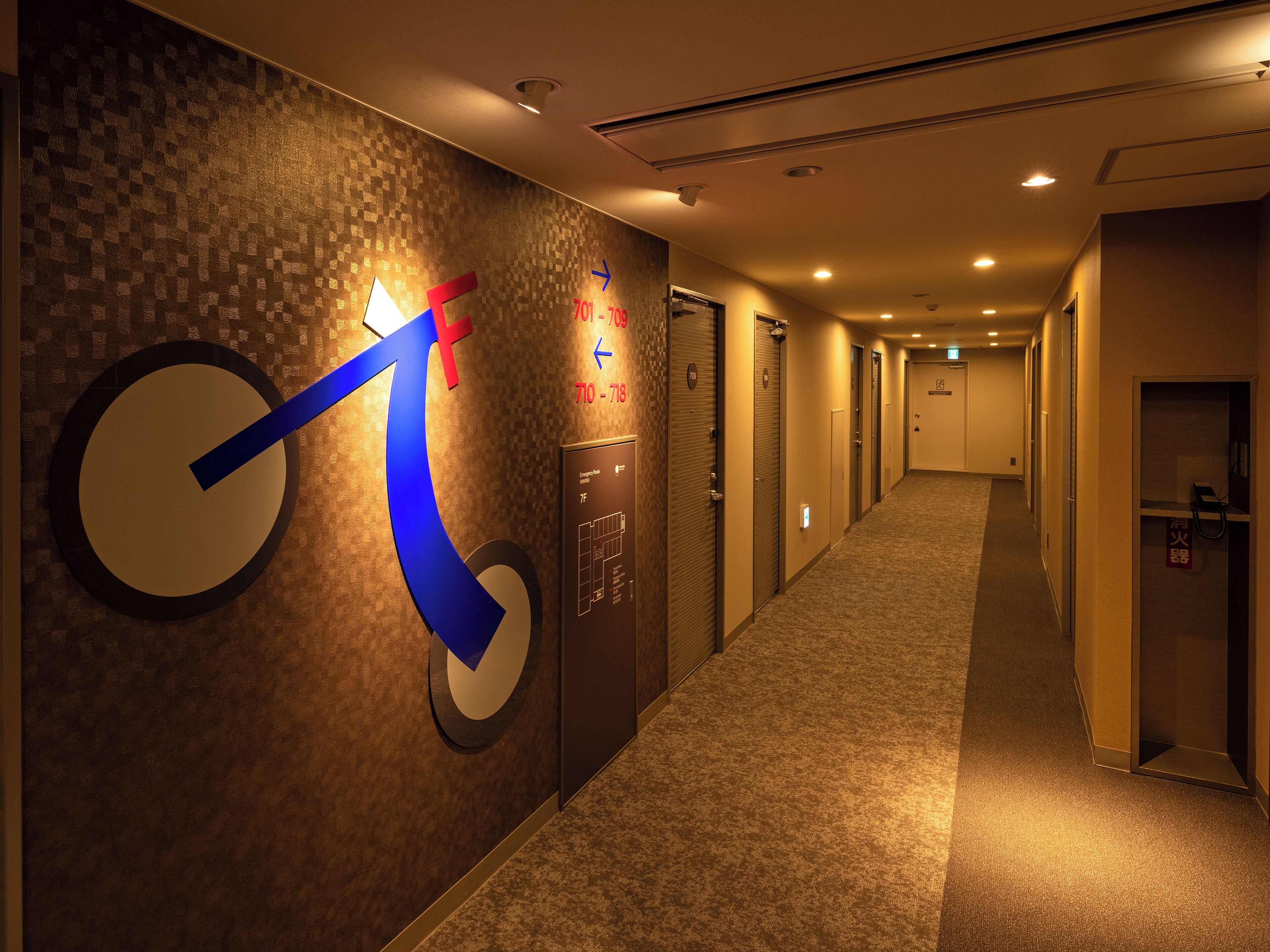 A hotel hallway with directional arrows on the wall, a carpeted floor, and doors leading to rooms.