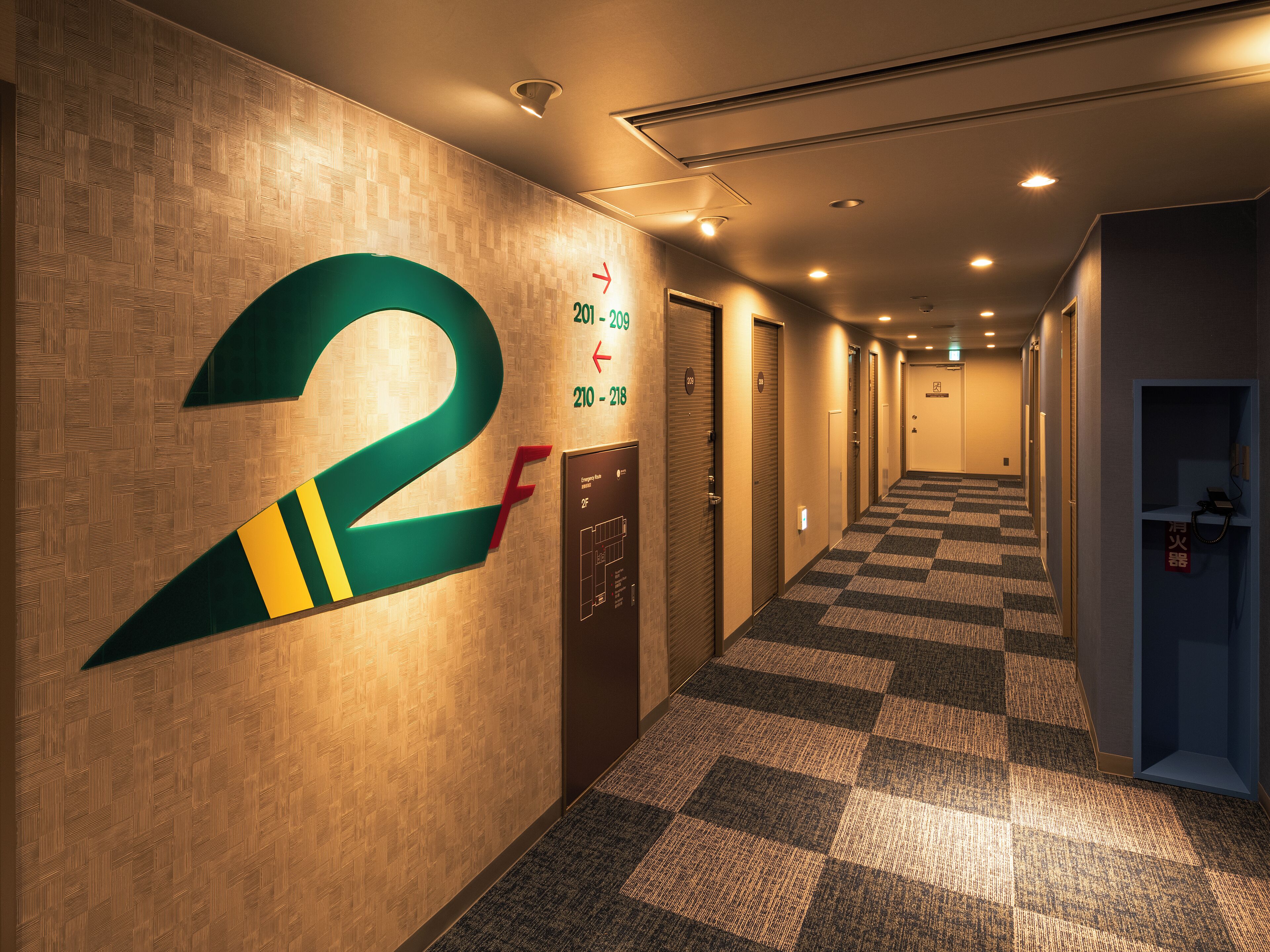 Un couloir d’hôtel avec un tapis à carreaux, des portes numérotées et une grande enseigne « 2F » sur le mur.