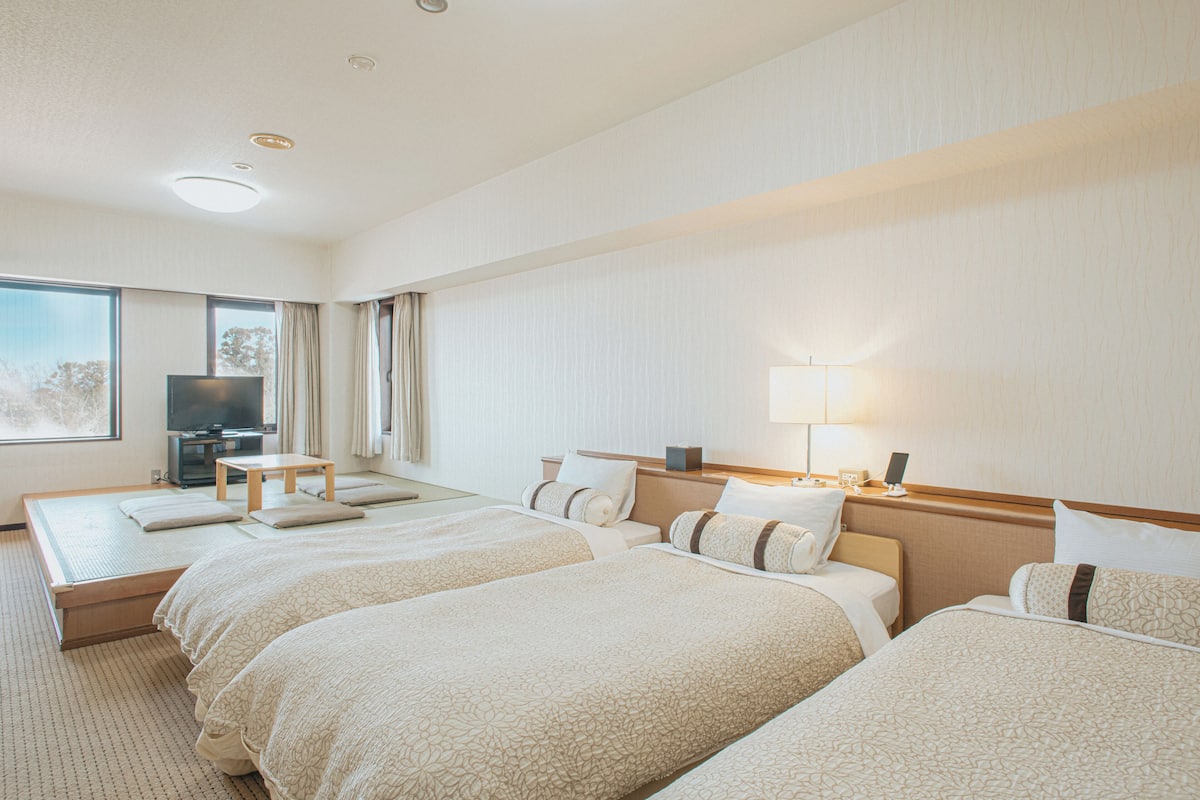 A hotel room with three beds, a television, a bedside table, and a lamp.