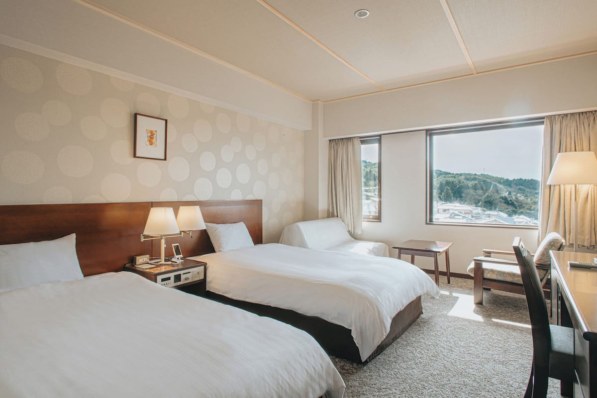 A hotel room with two beds, a desk, a chair, and a window with a view of a mountainous landscape.