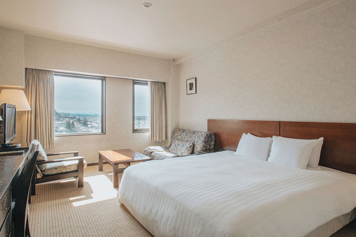 A hotel room with a large bed, a desk with a lamp, a chair, and a view of the city through the window.