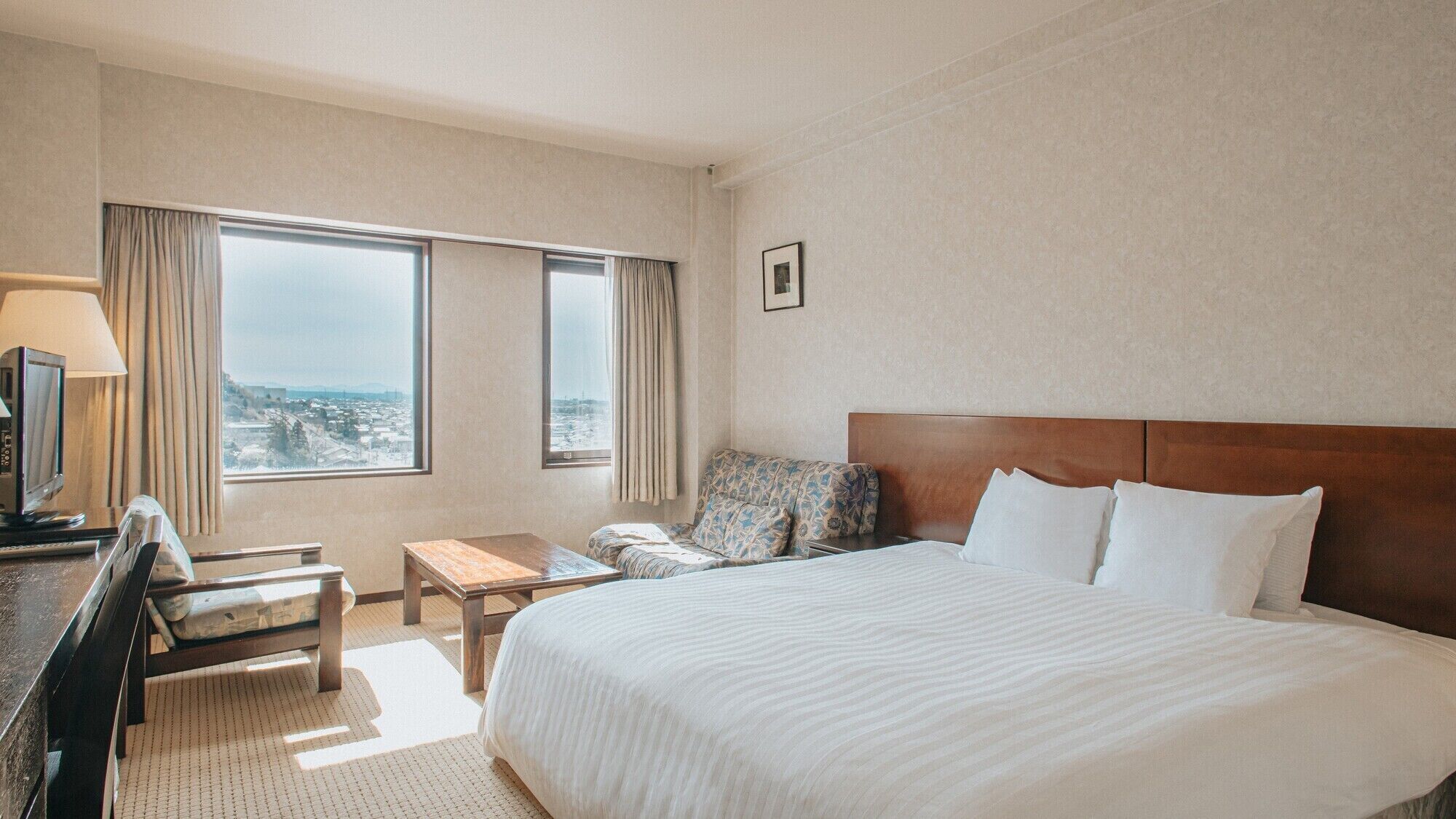 A hotel room with a large bed, a desk, a chair, a TV, and a view of the city through the windows.
