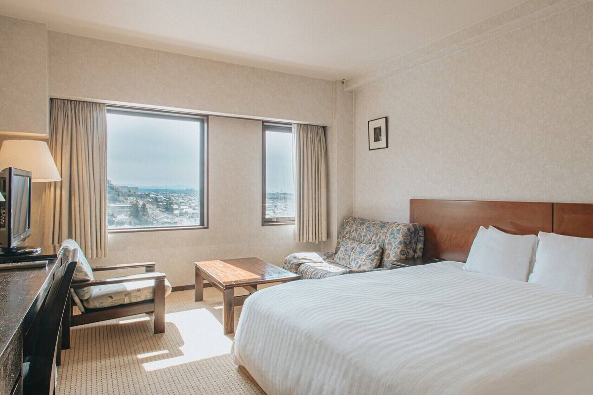 A hotel room with a large bed, a desk, a chair, a TV, and a view of the city through the windows.