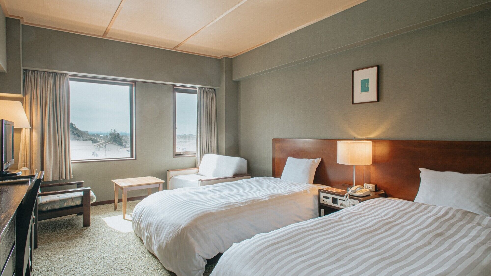 A hotel room with two beds, a desk, a chair, a bedside table, a lamp, and a view of the city.