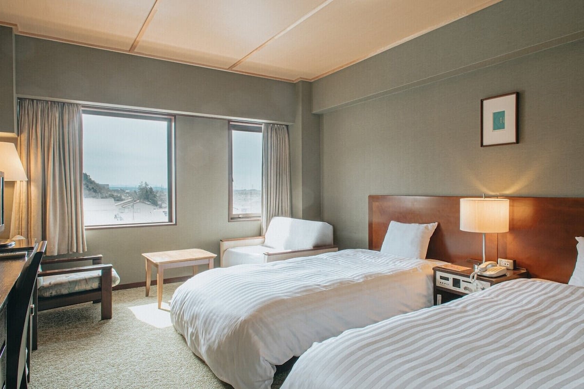 A hotel room with two beds, a desk, a chair, a bedside table, a lamp, and a view of the city.