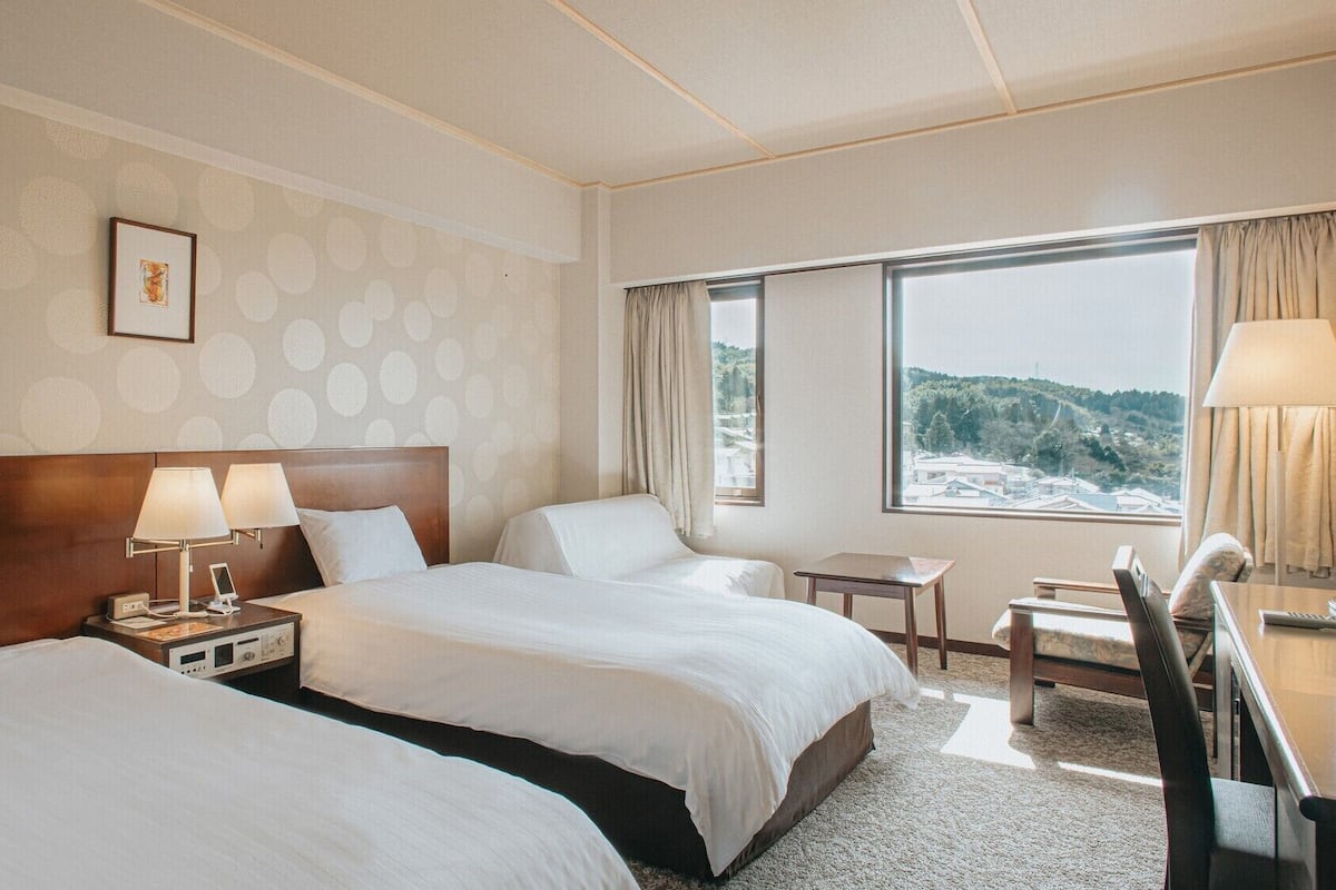A hotel room with two beds, a desk, a chair, a window with a view, and a framed picture on the wall.