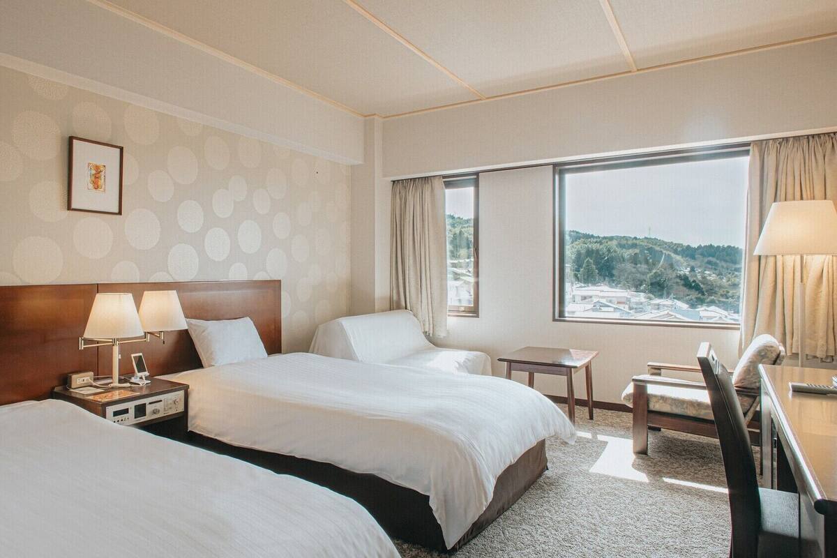 A hotel room with two beds, a desk, a chair, a window with a view, and a framed picture on the wall.