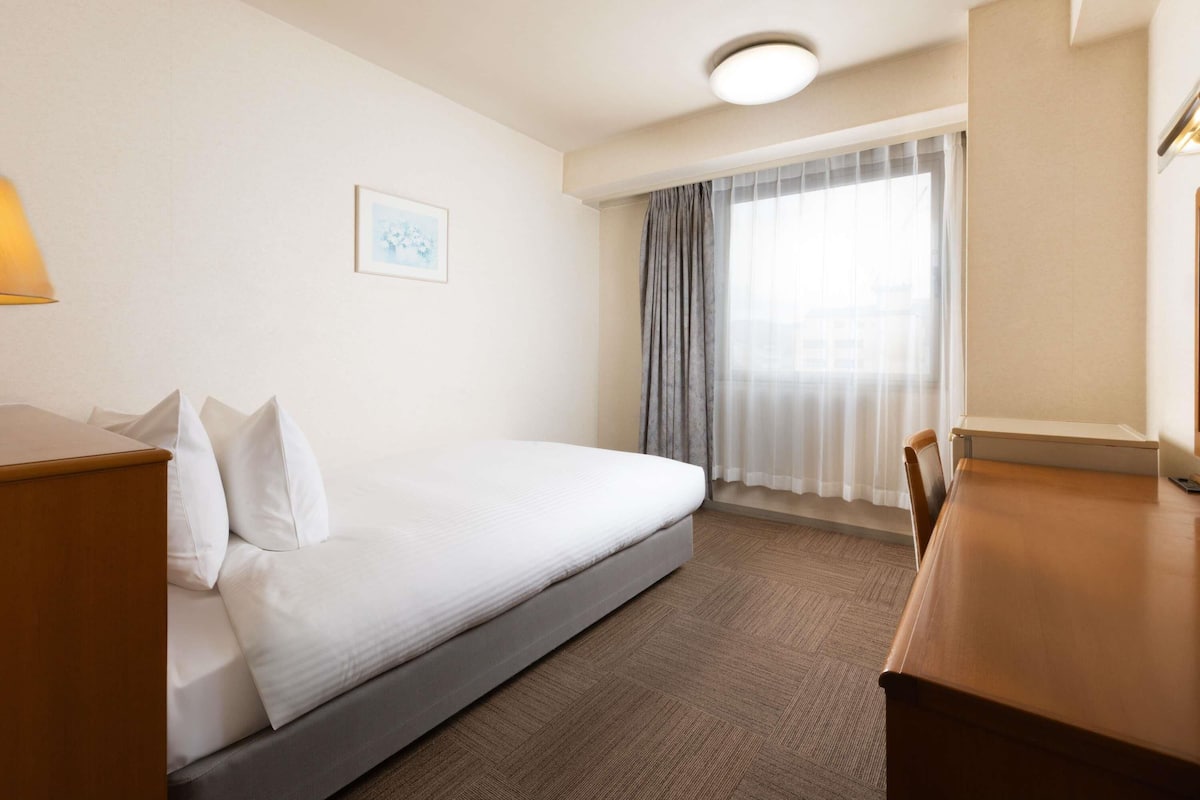 Ein Hotelzimmer mit Bett, Schreibtisch, Stuhl, Lampe, einem Vorhangfenster und einer Deckenleuchte.