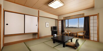Ein traditioneller japanischer Raum mit Tatami-Bodenbelag, einem niedrigen Tisch, zwei Stühlen, einem Fernseher und einer Schiebetür mit Blick auf die Stadt.