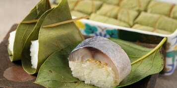 A traditional food item wrapped in leaves, possibly a type of rice dumpling, with a bowl of similar items in the background.