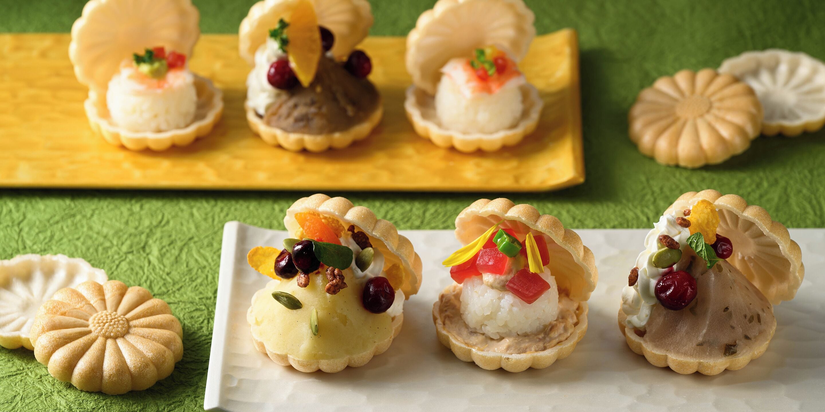 A variety of desserts in shell-shaped containers, some with fruits and nuts, on a green textured surface.