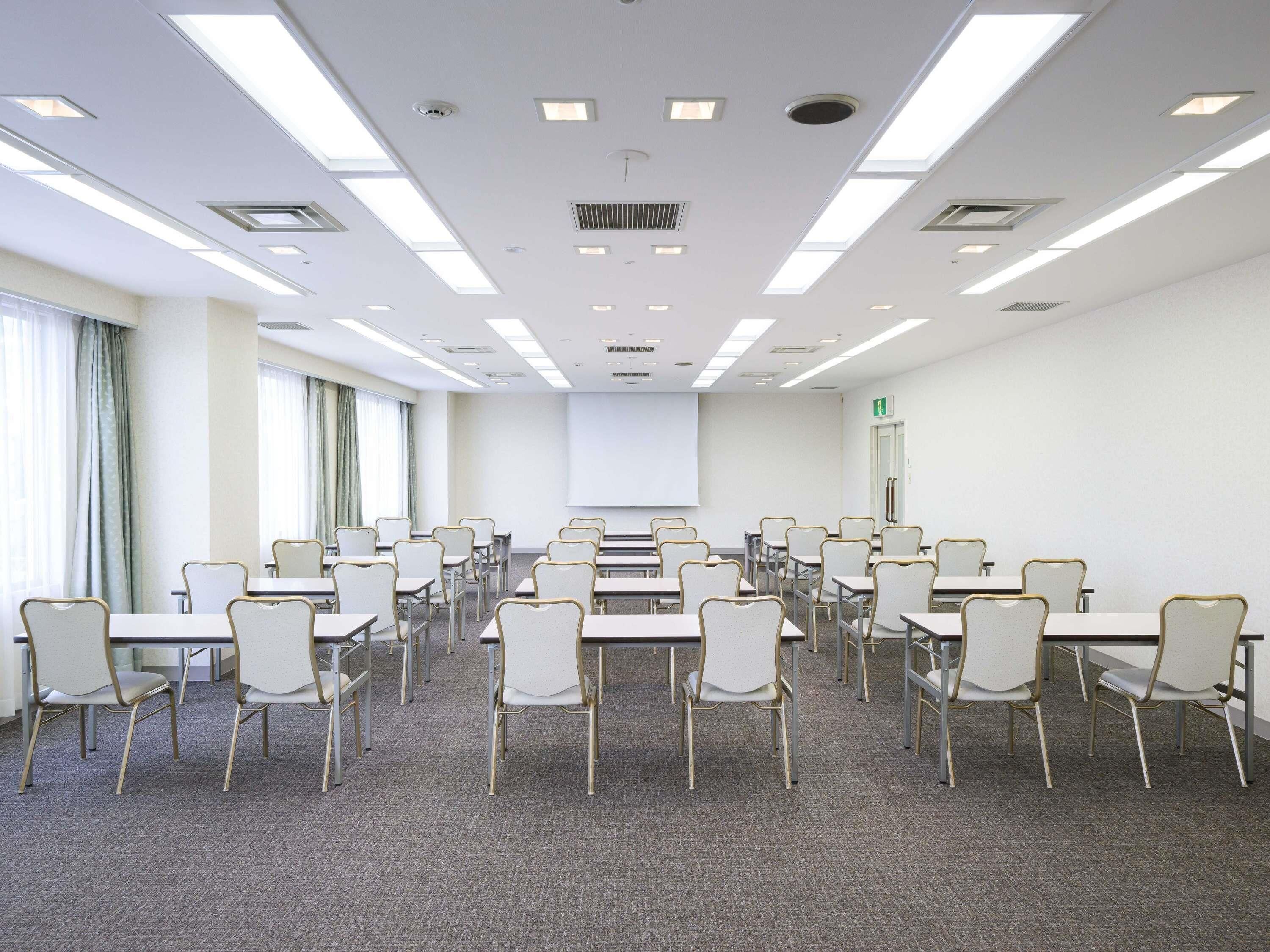 A conference room with rows of tables and chairs, a projector screen, and a door with an emergency exit sign.