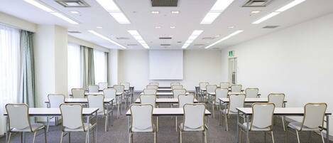 A conference room with rows of tables and chairs, a projector screen, and a door with an emergency exit sign.
