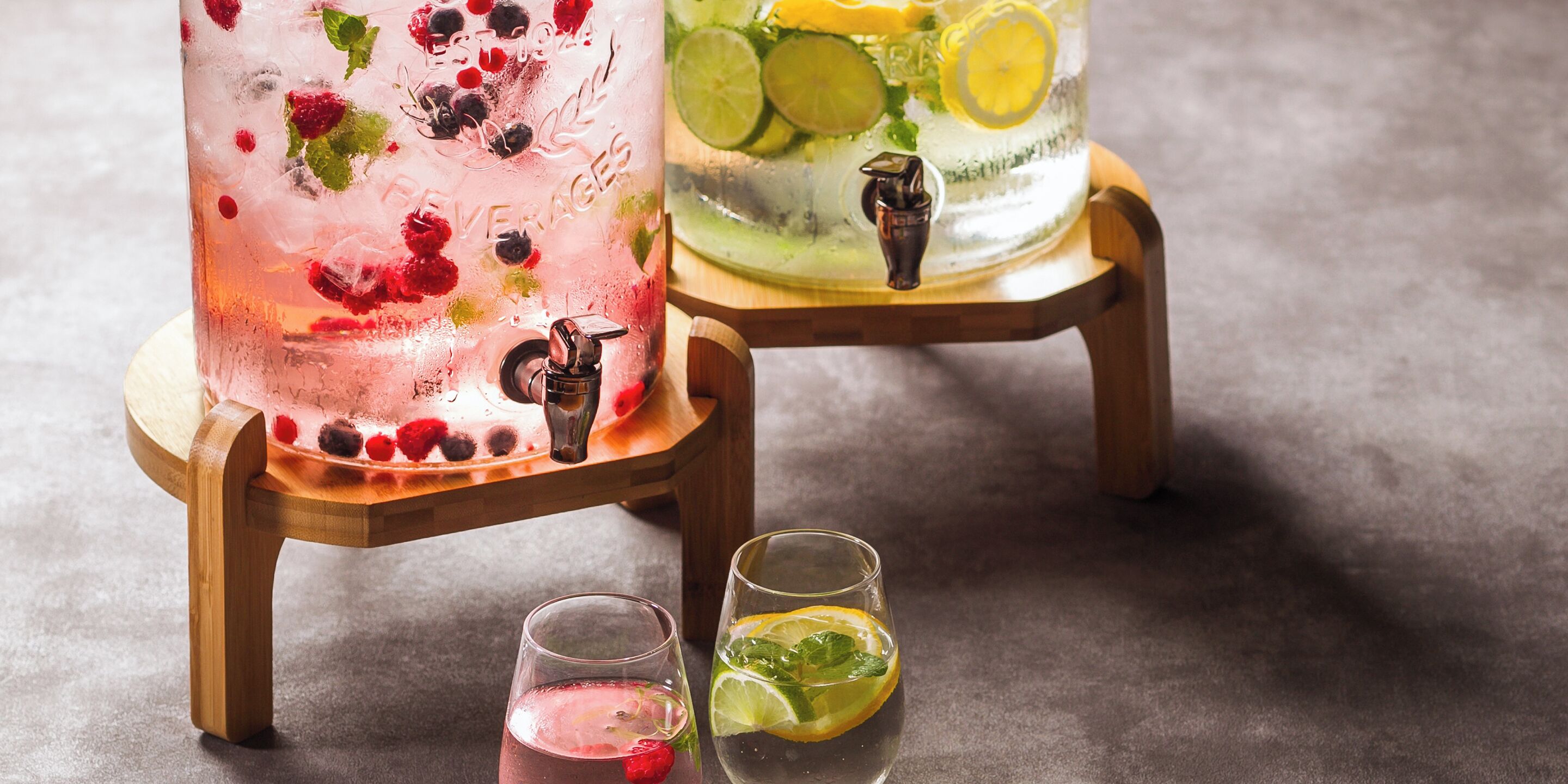 Two large glasses of iced beverages on wooden stands, one with berries and mint, the other with citrus slices.