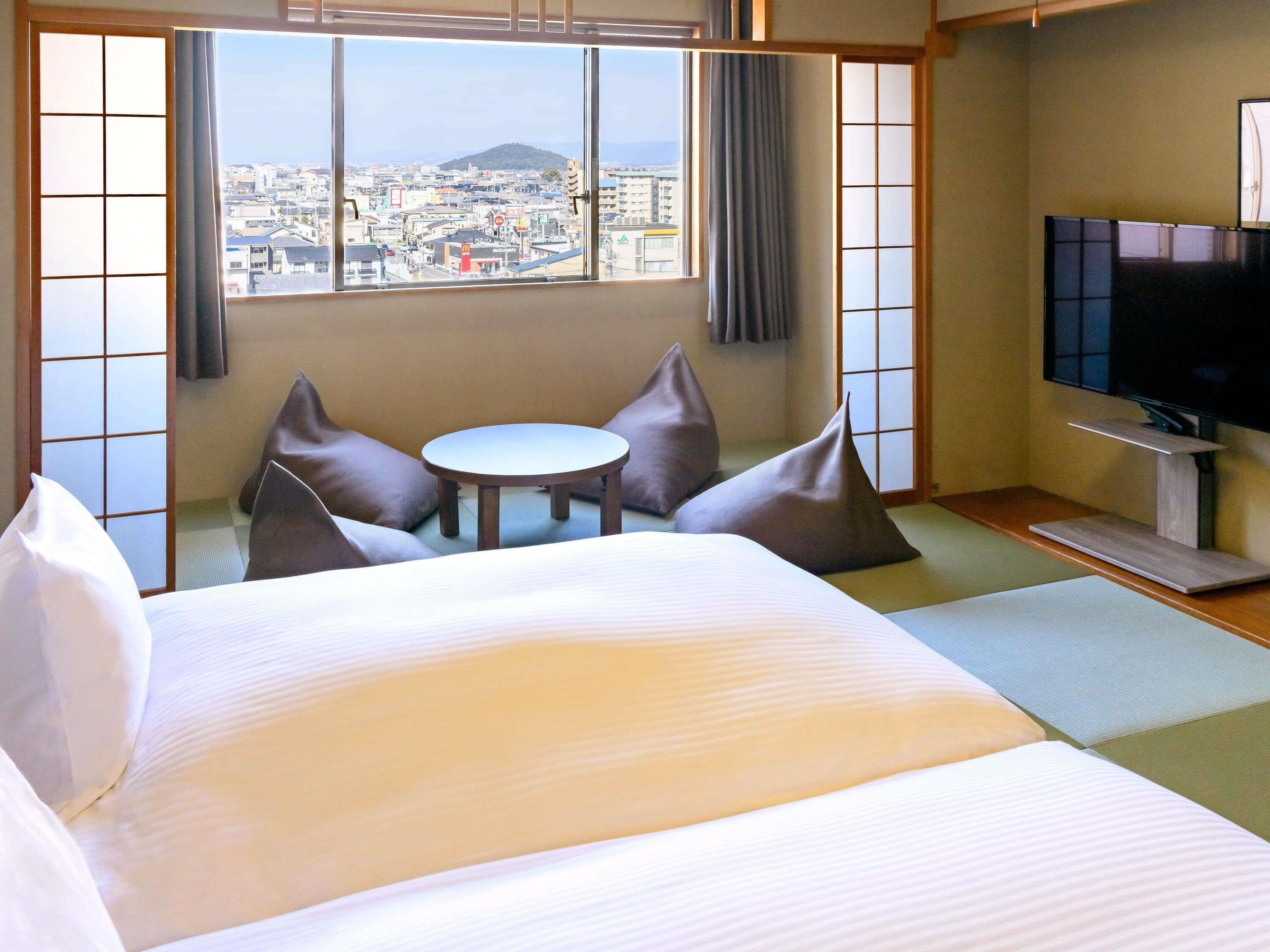 A hotel room with a bed, a small table, two chairs, a TV, and a view of a cityscape through large windows.