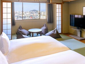 Habitación de hotel con cama, una mesita, dos sillas, televisión y vistas a un paisaje urbano a través de amplias ventanas.