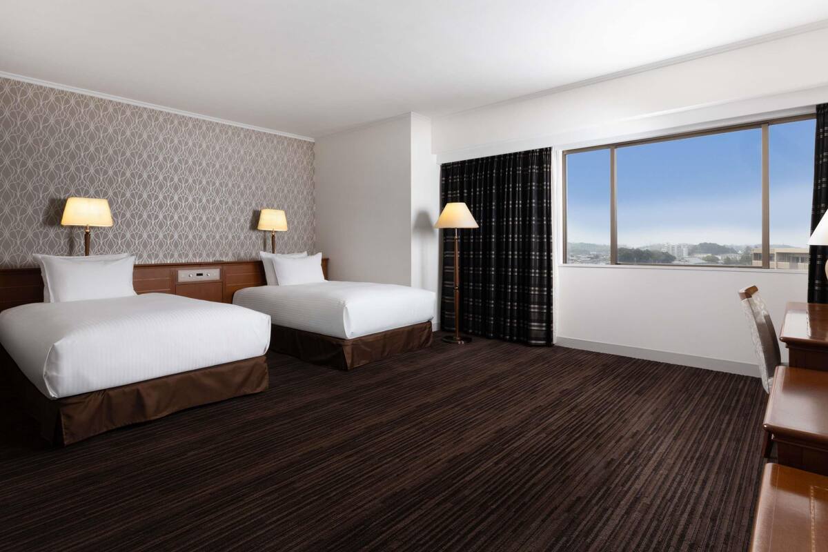 A hotel room with two beds, a desk, a chair, a window with a city view, and a patterned wall.