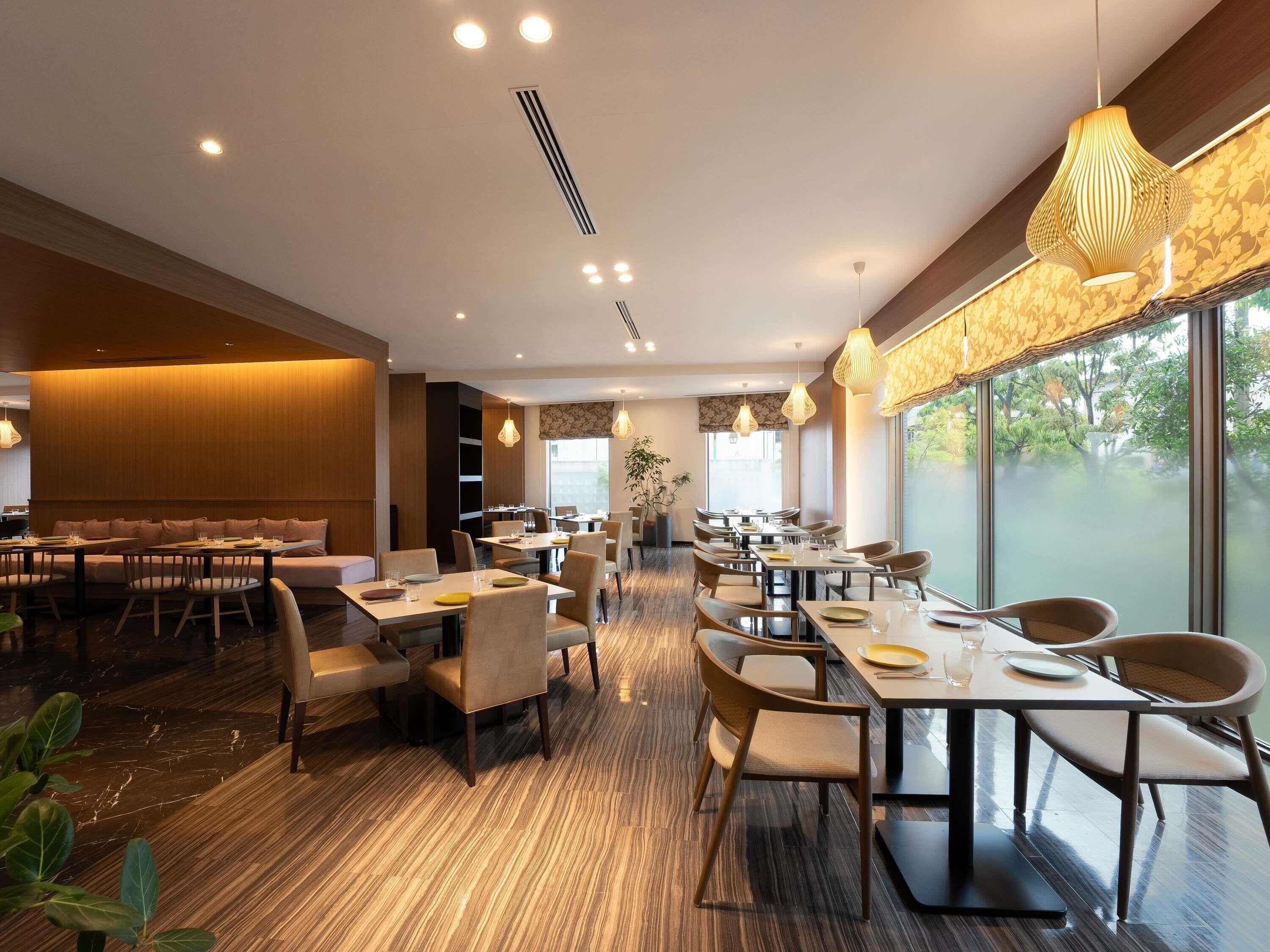 A modern dining area with multiple tables and chairs, a wooden floor, and large windows offering a view of greenery.