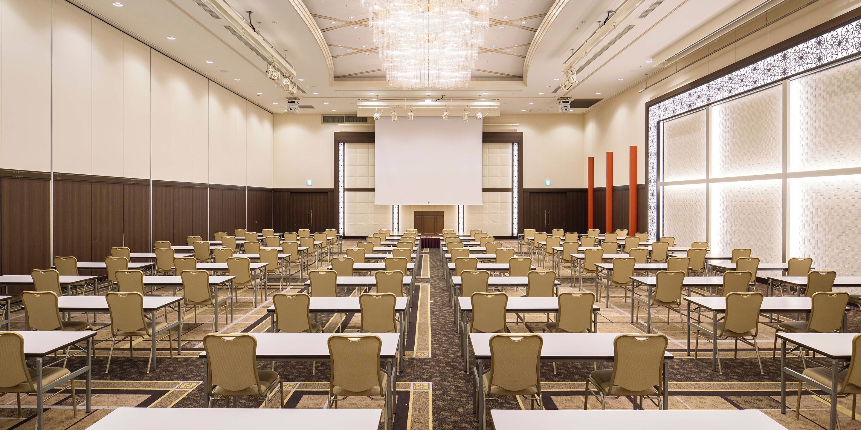 A conference room with rows of tables and chairs, a large screen at the front, and a patterned wall on one side.