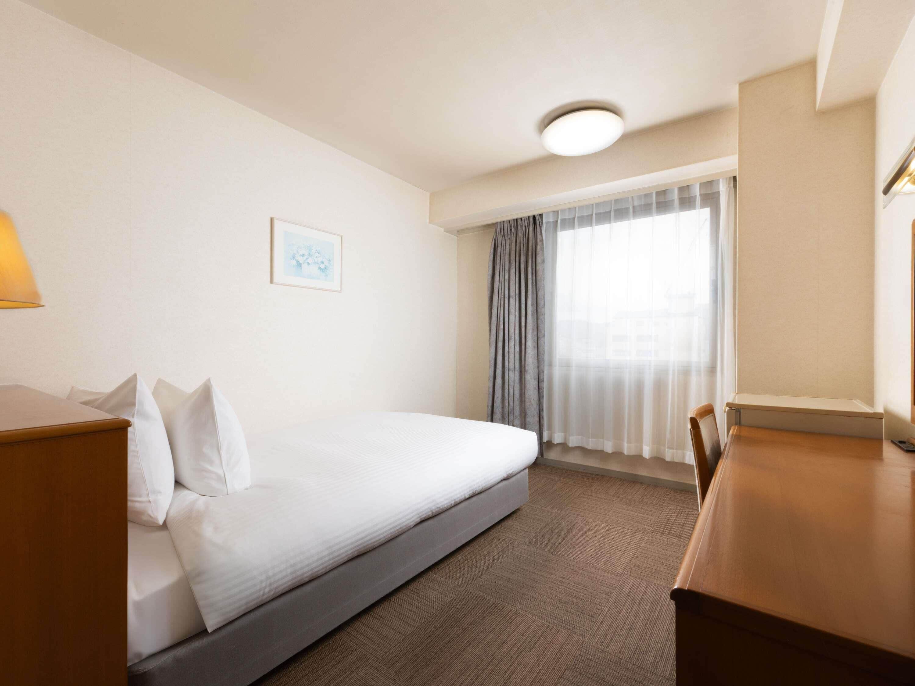 Habitación de hotel con cama, escritorio, silla, lámpara, ventana con cortinas e iluminación en el techo.