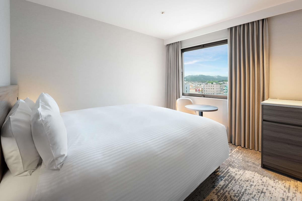 Ein Hotelzimmer mit einem großen Bett, einem kleinen Tisch, einem Sessel und Blick auf die Stadt durch das Fenster.