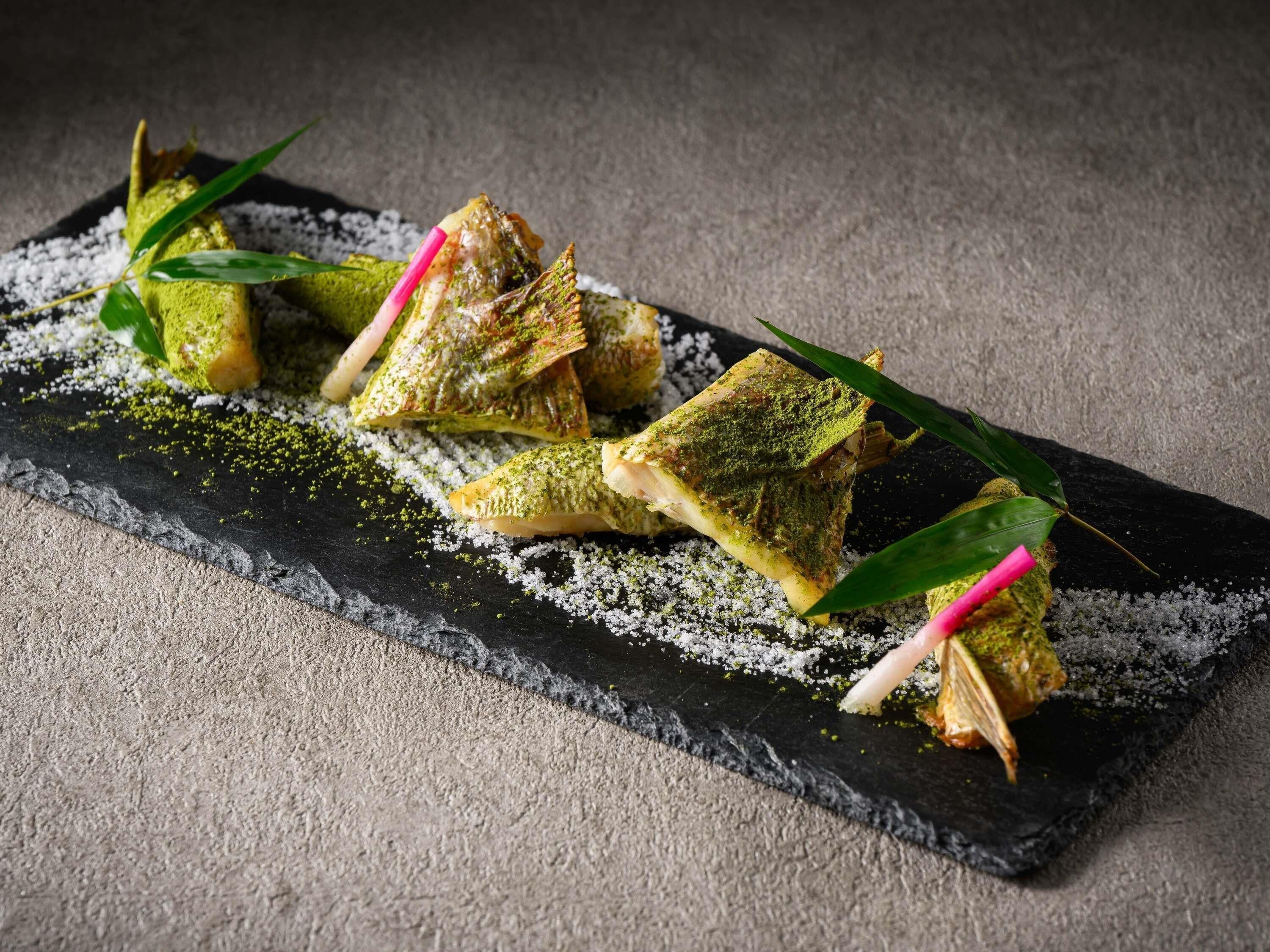 A dish with green and white powdered toppings, garnished with green leaves and pink sticks, on a black slate plate.