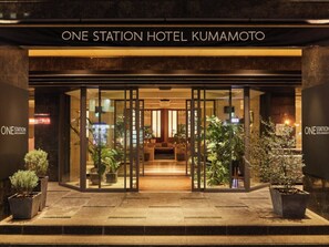 The entrance of the One Station Hotel Kumamoto with a modern design, featuring glass doors and potted plants.