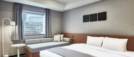 A modern hotel room with a large bed, a seating area with a sofa and pillows, a small round table, and a floor lamp.