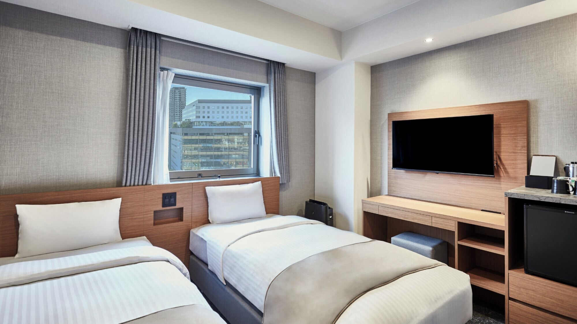 A hotel room with two beds, a TV, a desk, and a window with a city view.