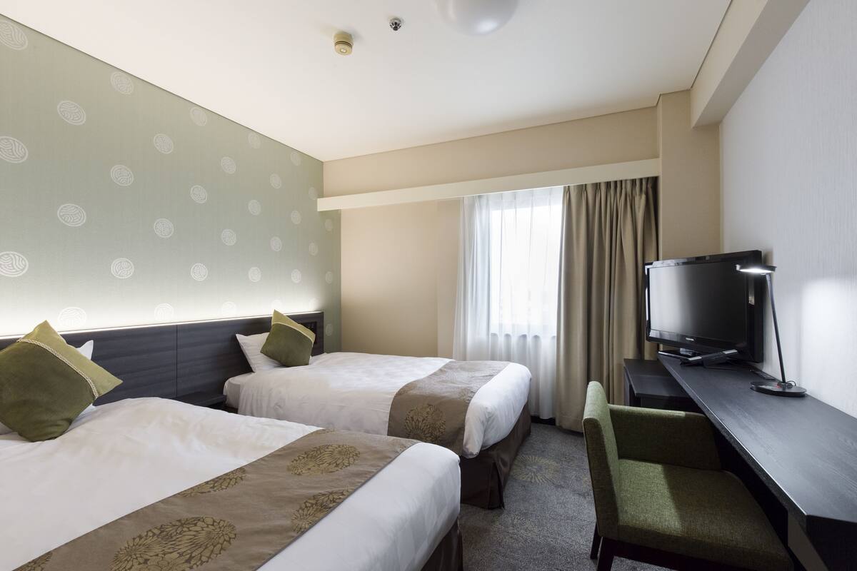 A hotel room with two beds, a desk, a television, and a window with curtains.