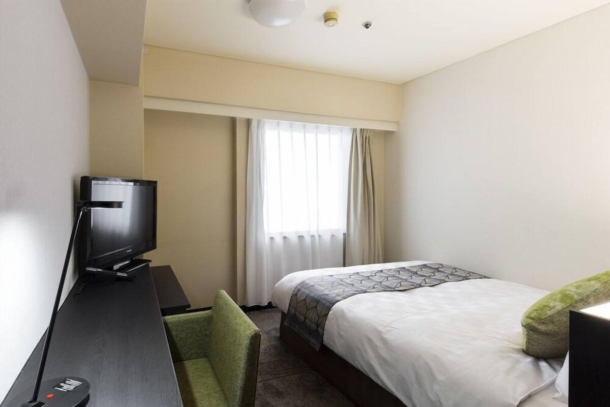 A hotel room with a bed, a desk with a television, a chair, and a window with curtains.