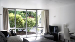 A modern hotel room with a large sliding glass door to a garden, a bed with a grey and white striped blanket, a desk with a laptop, and a TV. - Pacific Park (Dunedin)