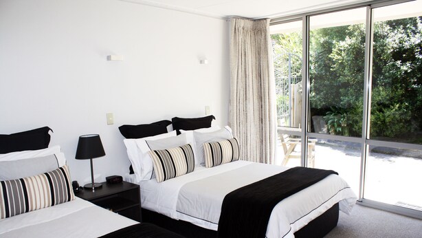 A hotel room with two beds, a nightstand with a lamp, and a large window offering a view of greenery. - Pacific Park (Dunedin)
