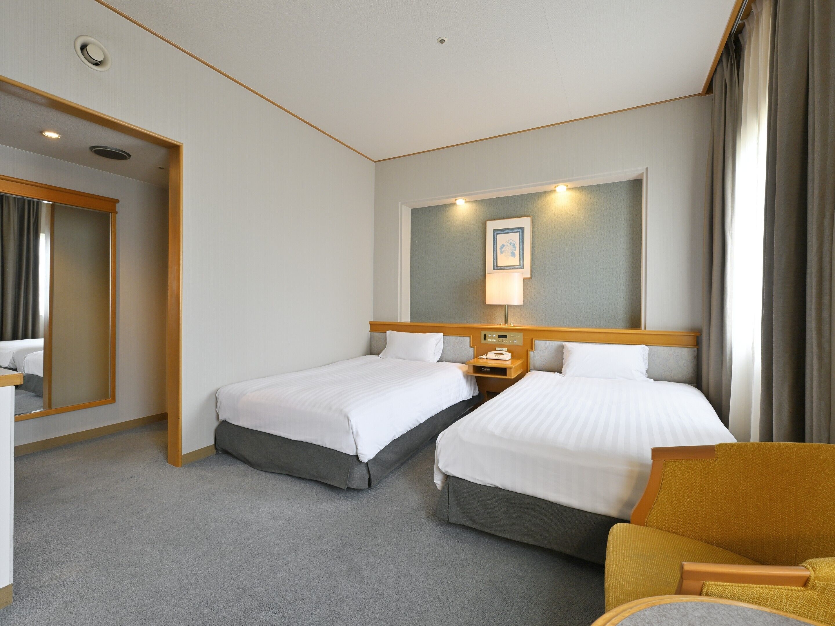 Habitación de hotel con dos camas, una mesita de noche con lámpara, un espejo grande y una silla.