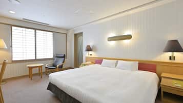 A hotel room with a large bed, a chair, a small table, and a desk with a lamp.