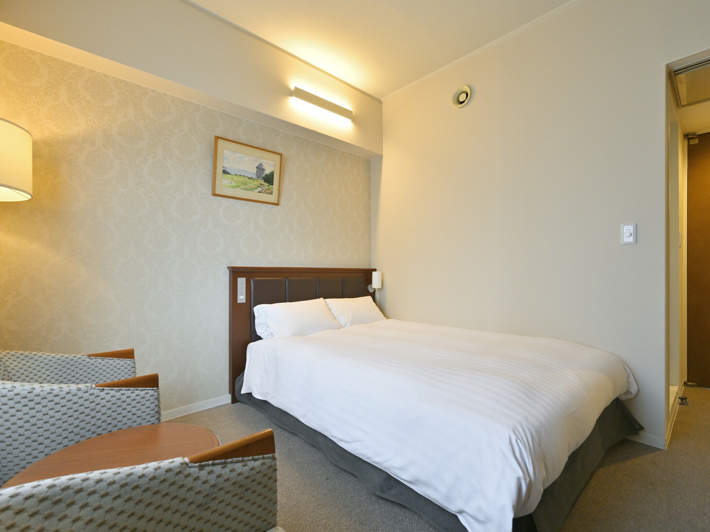 A hotel room with a bed, a desk, and a chair. There is a painting on the wall and a light fixture above the bed.