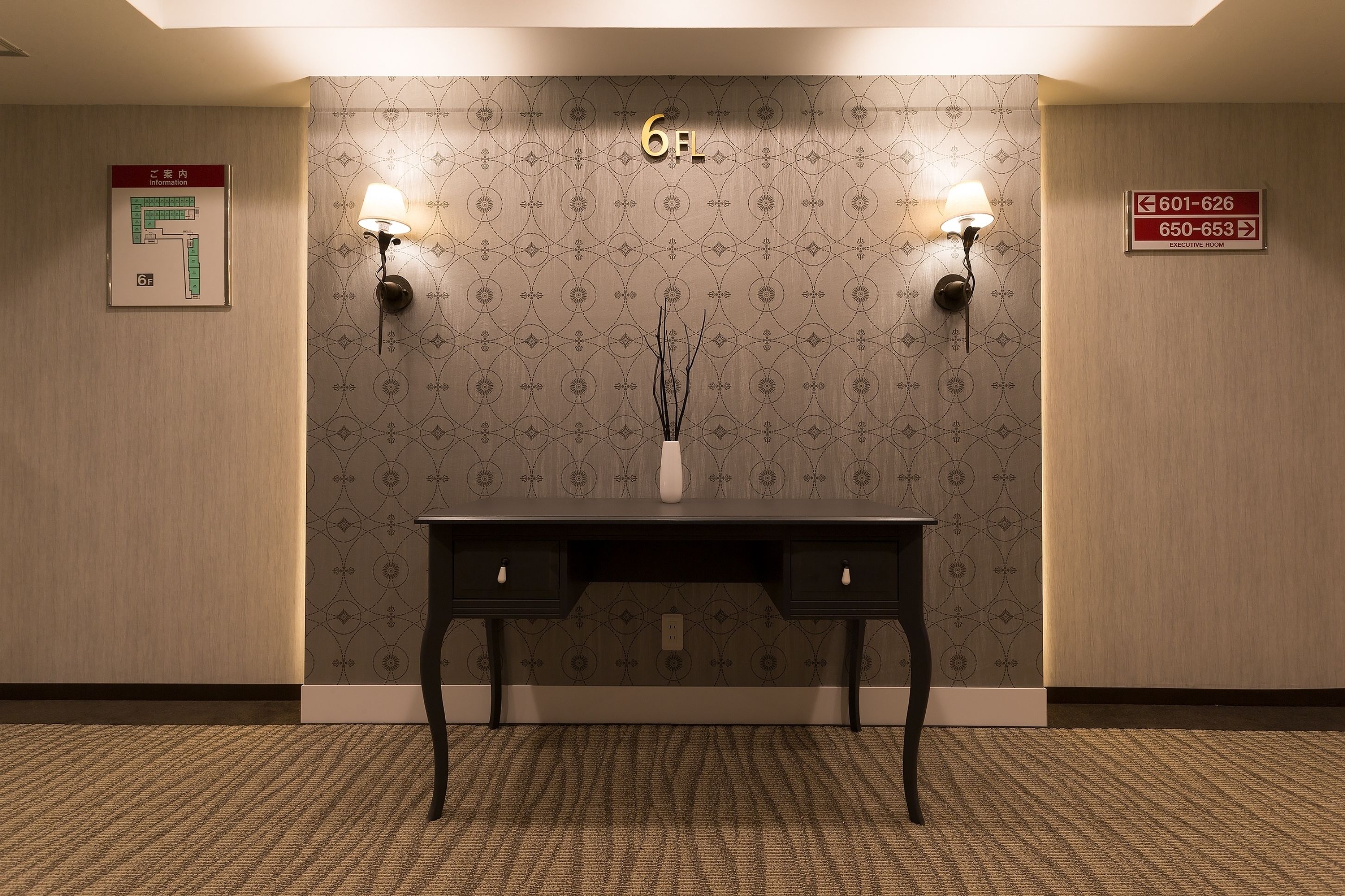 A hotel lobby with a reception desk, two wall-mounted lamps, a decorative vase, and directional signs for the executive room and a phone number.