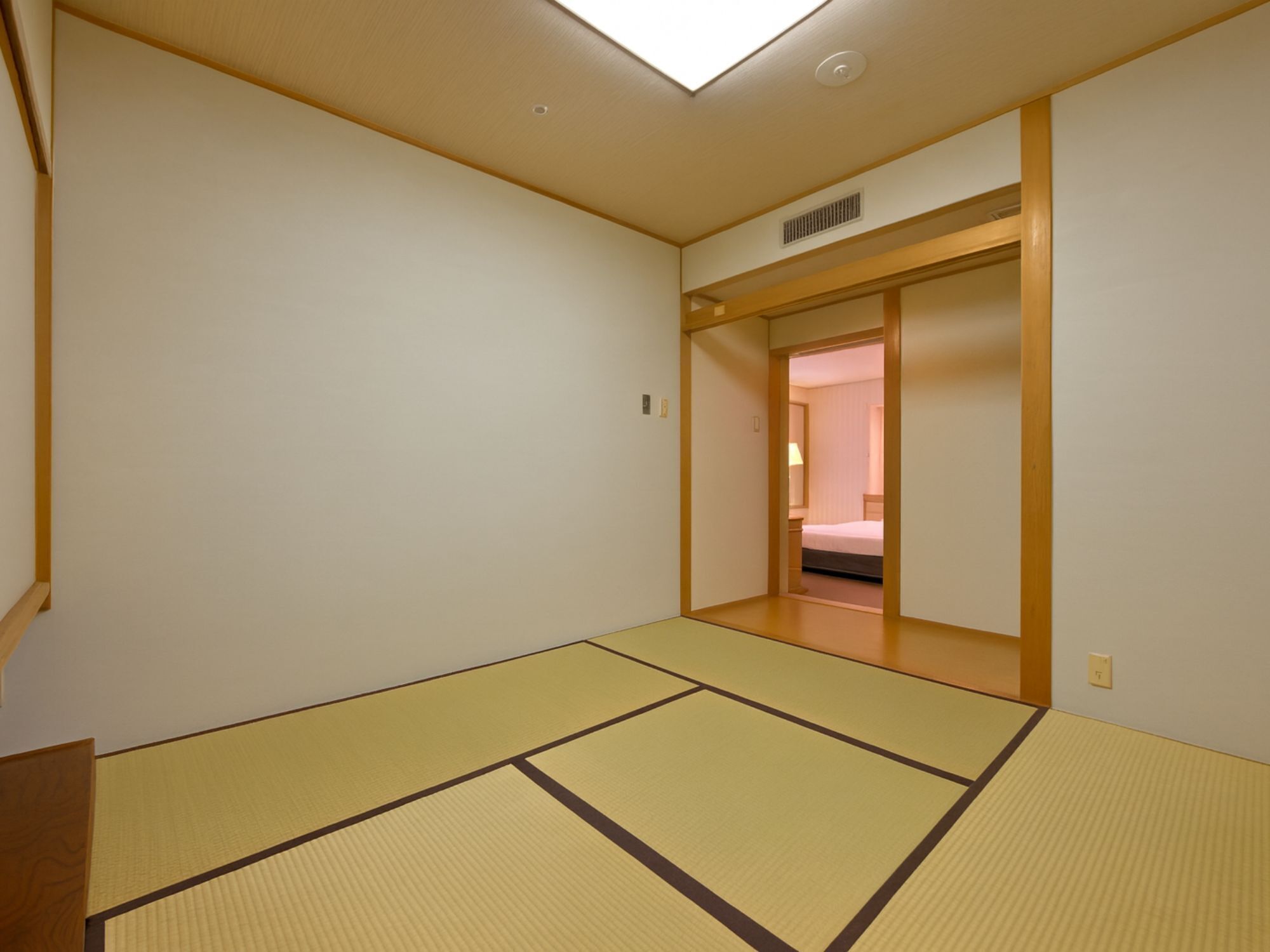 A room with tatami mats, a wooden door, and a bed in the adjacent room.