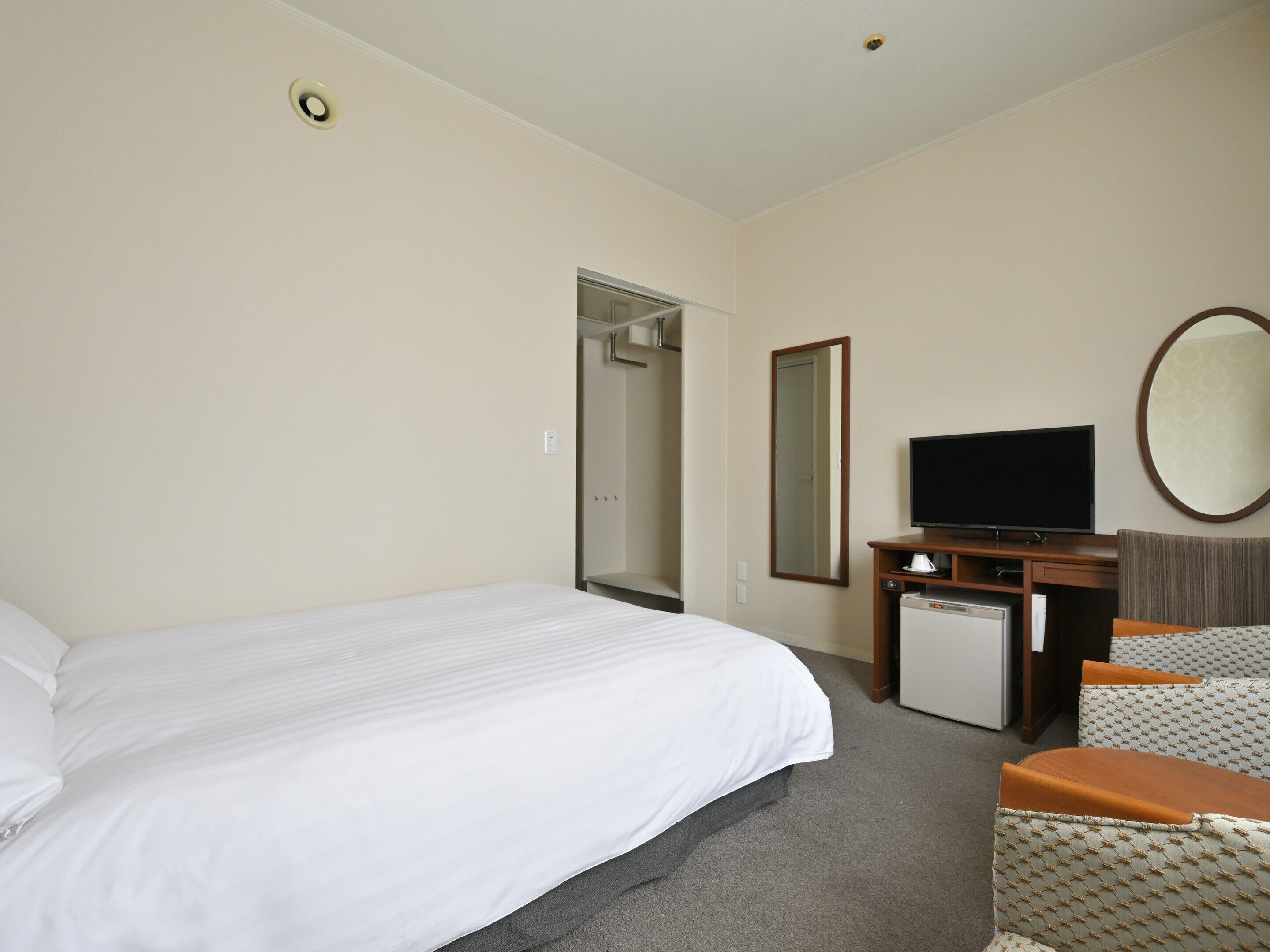 A hotel room with a bed, a television, a small refrigerator, a mirror, and a seating area with a cushioned chair.