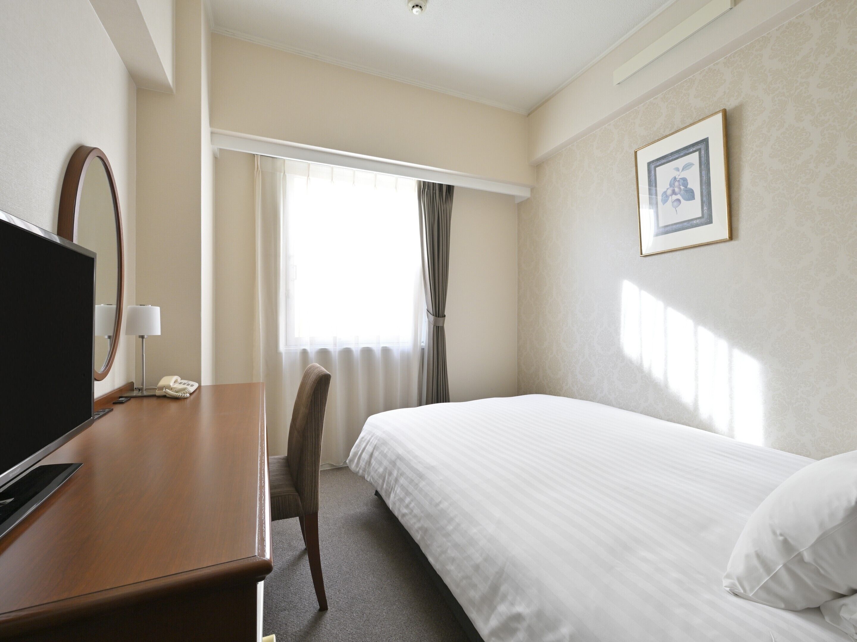 A hotel room with a bed, a desk with a TV, a chair, a window with curtains, and a framed picture on the wall.