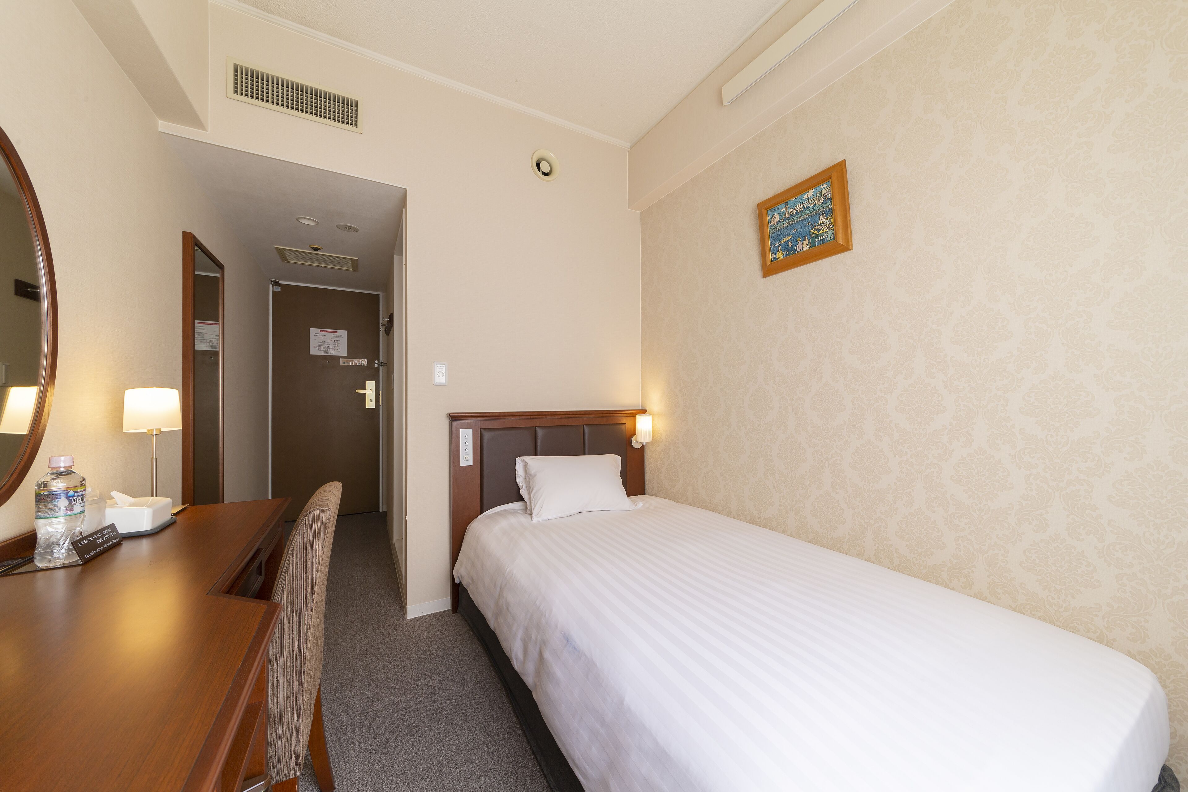 A hotel room with a single bed, a wooden desk with a chair, a mirror, and a wall-mounted light fixture.