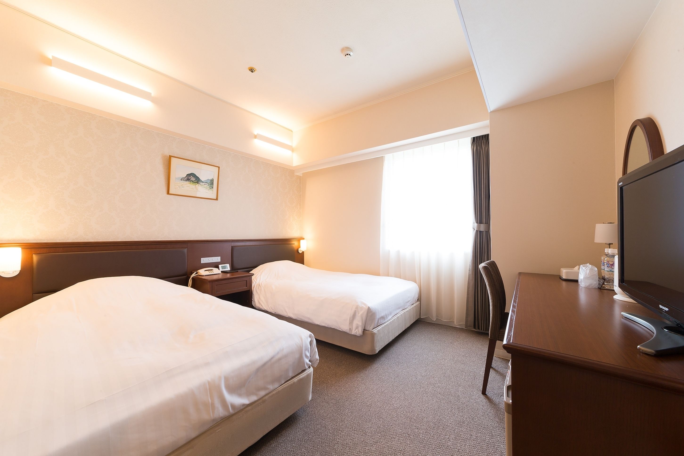 A hotel room with two beds, a desk with a computer, a TV, and a window with curtains.