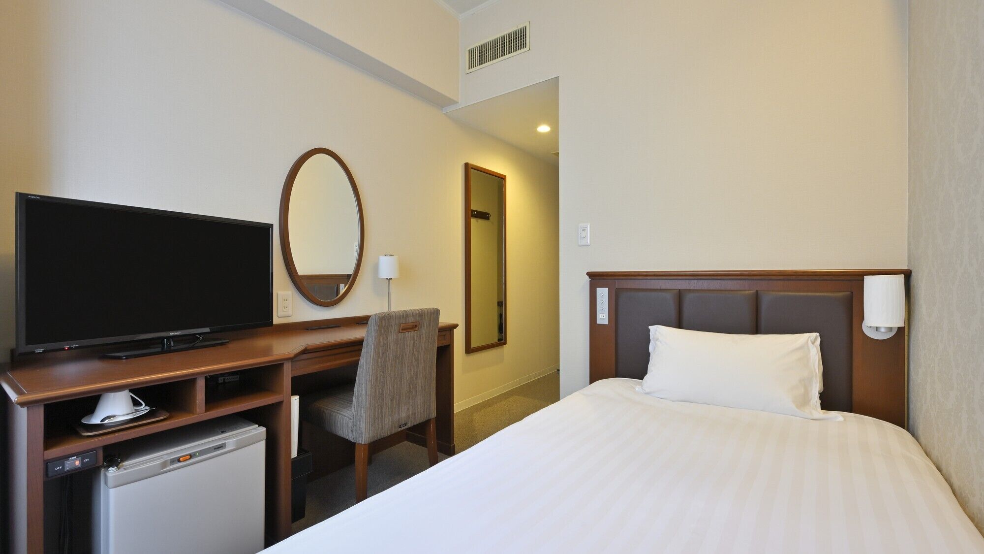 A hotel room with a bed, a TV, a desk with a chair, a mirror, and a small refrigerator.
