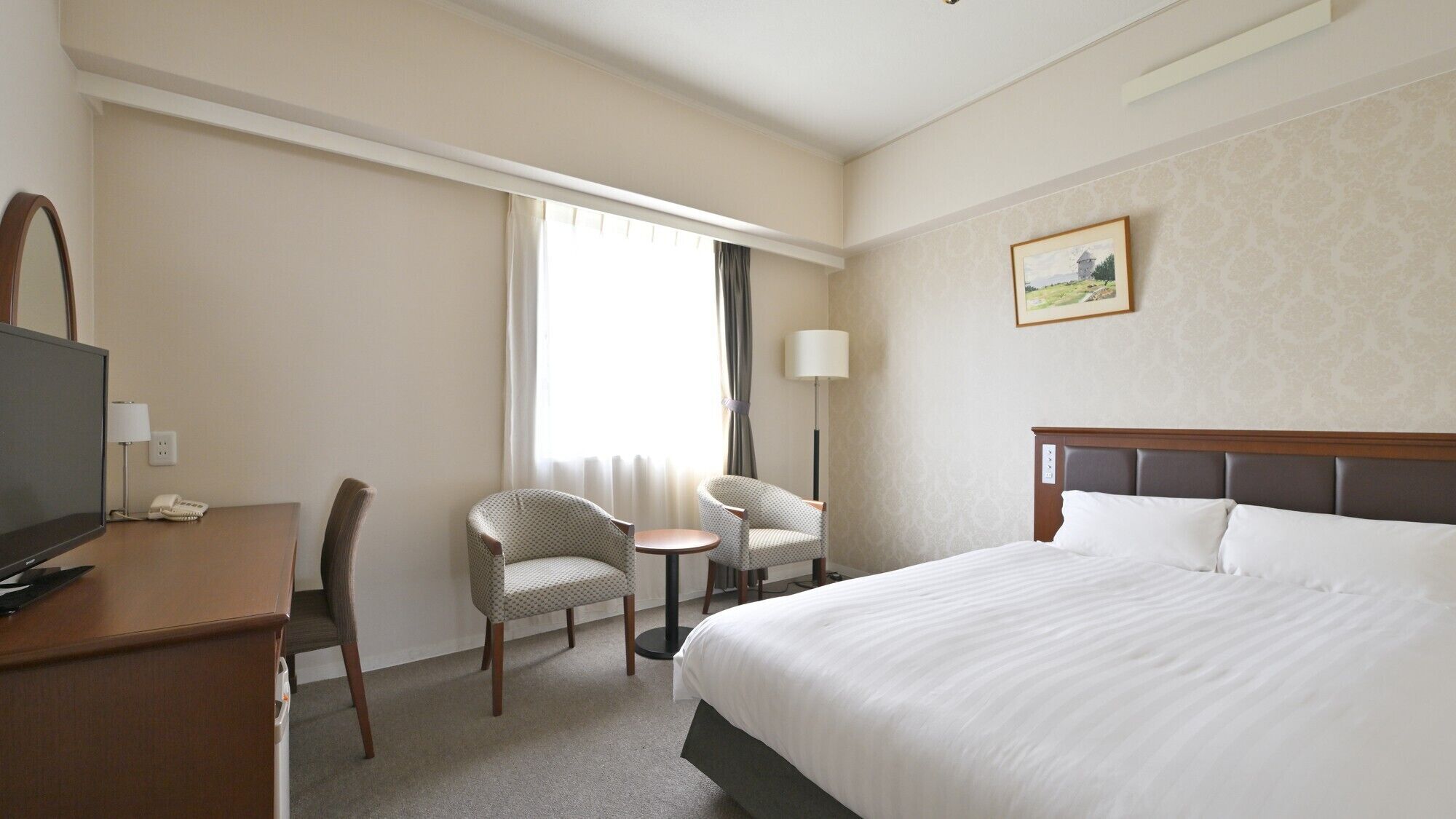 A hotel room with a bed, a desk with a television, two chairs, a small table, a lamp, and a painting on the wall.