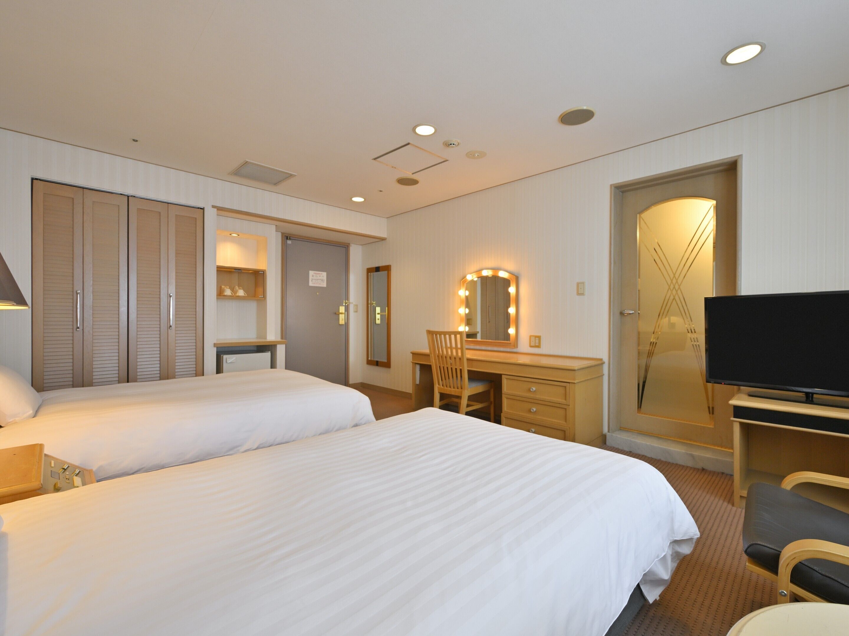 Habitación de hotel con dos camas, un escritorio con silla, un televisor y un espejo con luces.