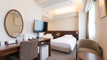A hotel room with a bed, a desk with a computer, a chair, a small table, and a TV.